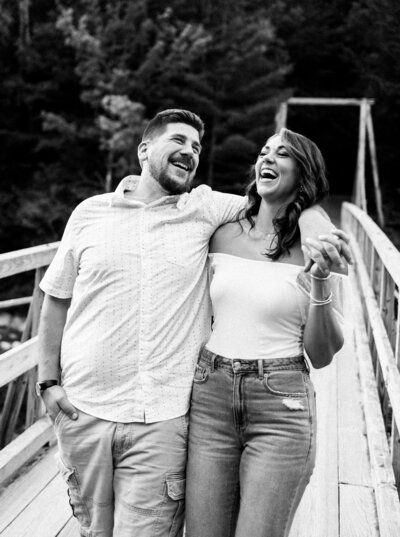 Groom slings arm over bride's shoulder while laughing during engagement session in Lincoln, New Hampshire