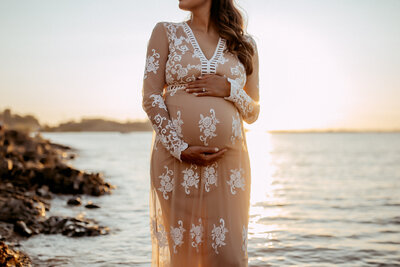 An expecting mother in a sheer lace dress holding her bump at the lake while the sun sets behind her. 