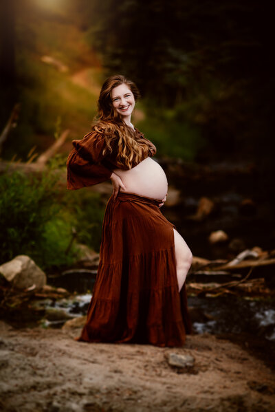 An expecting mother in a brown two piece standing by a stream in Estes Park Colorado at sunset. 