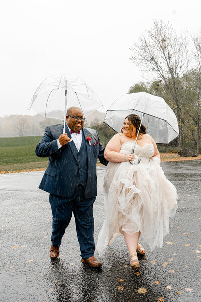 Bride and groom standing under a clear umbrella during a rainy wedding day, sharing a candid romantic moment.