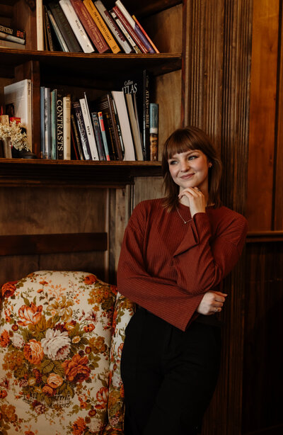 A woman in a a maroon top poses in a coffee shop in Essex, Vermont. She stands stoically next to an elegant floral chair and bookshelf. A perfect coffee shot setting for capturing moments by a New England Portrait Photographer.