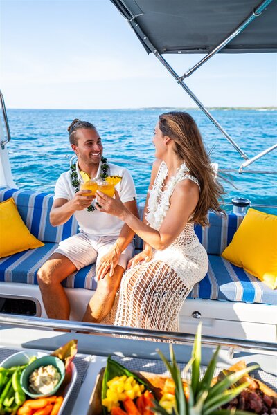 Engaged couple relaxing on a sailboat in Hawaii, smiling and clinking tropical drinks while sitting on blue and white striped cushions with ocean views.