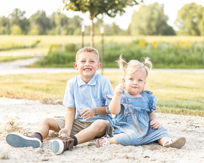 brother and sister sitting on the ground at a wildlife sanctuary, brother is wearing a blue shirt, khaki shorts smiling at the camera, sister has blond hair wearing a blue dress 