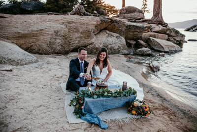 A couple enjoys a romantic Lake Tahoe elopement, sitting on a blanket by the sandy shore, toasting drinks as the sun sets. The woman wears a blue and white striped dress, and the man wears a white shirt. Snow-capped mountains and the lake’s clear waters create a stunning backdrop.