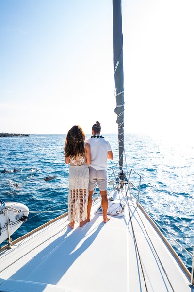 Couple standing on the front of a sailboat in Hawaii watching dolphins swim near the boat in bright, clear blue water.