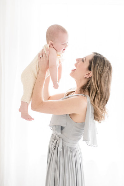 Austin Newborn Photographer Rebecca Dyan headshot sitting in white studio on cream chair wearing a floral dress smiling at the camera
