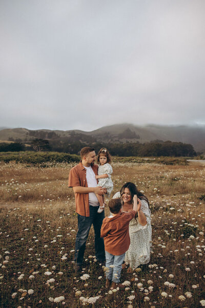 A family of five, including a mother, father, two girls, and a boy, standing in a flower-filled meadow with foggy hills in the background. the mother adjusts a young girl's hair.