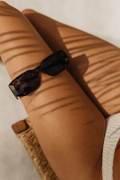 Close-up of a person lounging outdoors with smooth, tan skin, wearing a white swimsuit. Sunglasses rest on their thigh while soft shadows from nearby objects create intricate patterns on the skin.