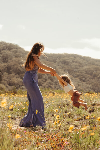 A woman in a blue jumpsuit swings a child by the arms in a field of wildflowers with hills in the background.
