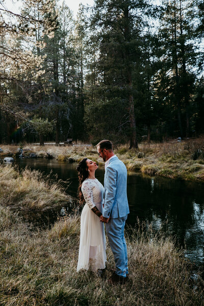 Bride and groom sharing a tender moment by a serene stream, surrounded by tall trees during their elopement