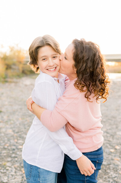 Mom in pink sweater squeezing her son while giving him a kiss on the cheek