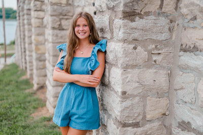 Girl wearing blue romper leans against grey brick building while smiling at the camera.