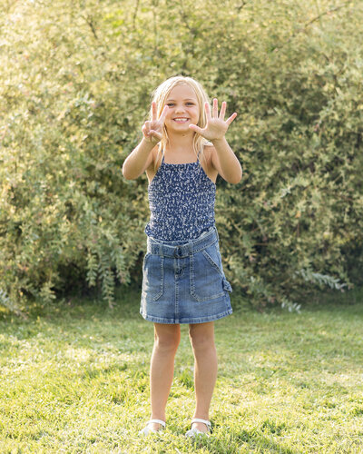 young girl standing in the grass in front of a bush, with seven fingers up smiling at the camera, wearing a sleeveless shirt and jean skirt