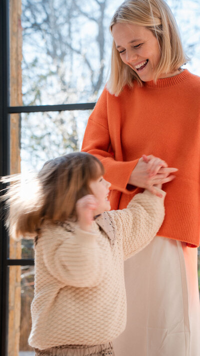 A joyful moment shared between parent and child dancing together, with orange and cream sweaters by a window.