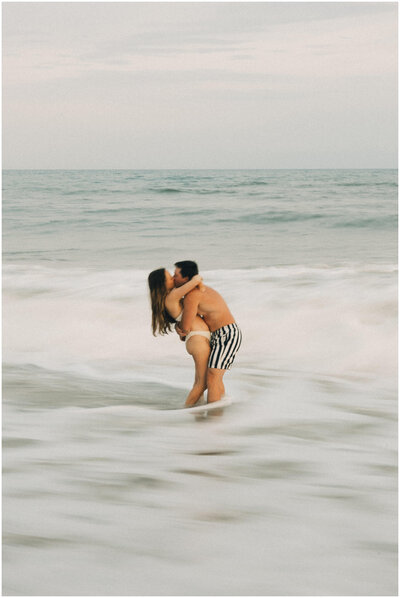 An engaged couple shares a passionate kiss in bathing suits in the water on the beach