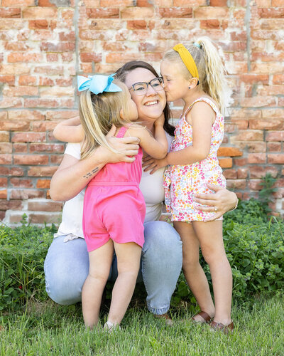 a mom knelt down with her two young daughters on each side giving her a kiss as she's smiling, they are standing in grass with a brick wall behind them