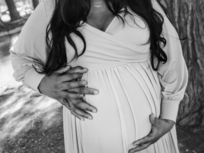 A black-and-white photo of a pregnant mother with long dark hair holding the top and bottom of her belly while her partner stands behind her and puts his hand on top of hers. By SAVI Photography - Photographer in Riverside