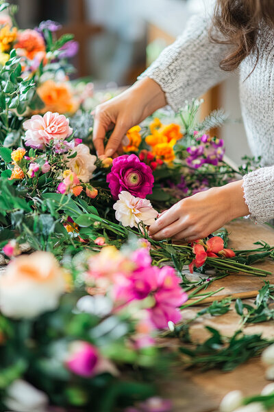 Branding image of a close up of a woman's hands preparing a wedding floral display with vibrant flowers.