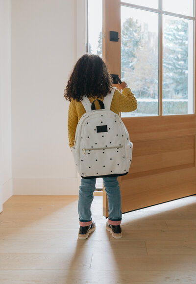 A young girl wearing a yellow sweater and polka dot backpack opens the front door to head outside on a winter day – Family Autism Support Coach at Chantal Hewitt Coaching