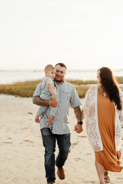 a couple walks hand in hand on the beach holding their son