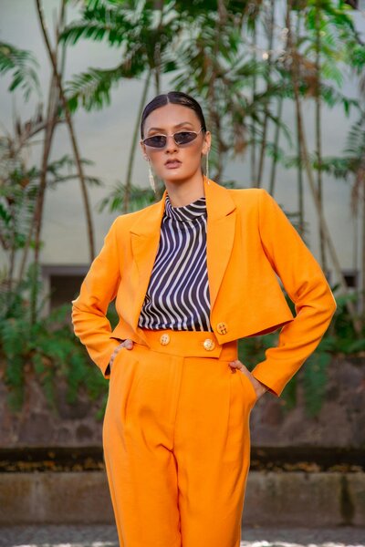 Image of a woman outside with an orange pant suit, blue and white striped shirt, and sunglasses looking powerful.