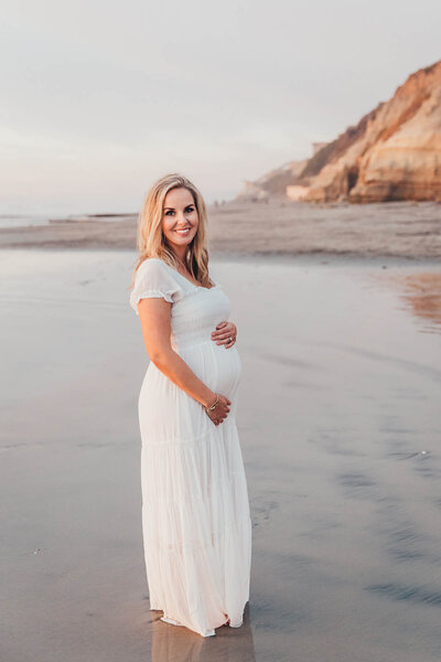 a blonde woman smiles at the camera holding her baby bump during her maternity photo session