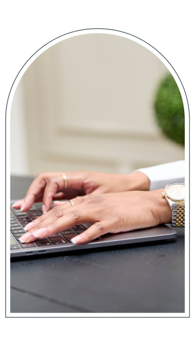  Close-up of a woman’s hands typing on a laptop keyboard, wearing gold rings and a watch, seated at a modern black desk.