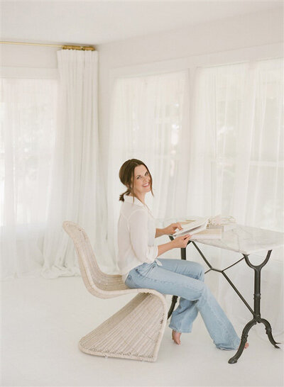 Meredith Maag smiles while seated at her desk in a natural light studio, working in jeans and a white blouse, reflecting her warm Kansas City brand.