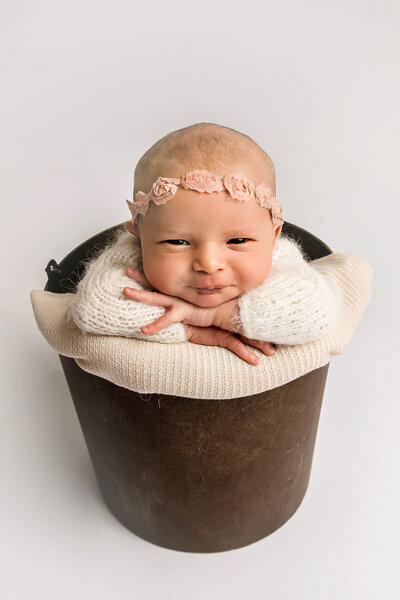 Connecticut newborn photographer captures adorable wide-awake smiling baby girl with pink floral headband posed in dark metal bucket with cream wraps