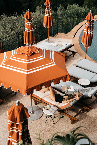 A person lounges on a poolside chair under an orange and white striped umbrella, reading a book. Nearby, other lounge chairs and umbrellas are set up by the pool. A towel drapes over a nearby surface, and trees are visible in the background.