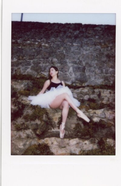 Michelle, dressed in her white ballerina tutu and point shoes with a black leotard, sits on a steep incline of rocks in San Francisco. Photographed by Renata Voci on Instax film.