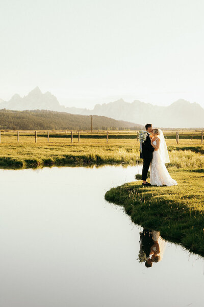 bride and groom standing next to a small pond embracing with mountains in the distance