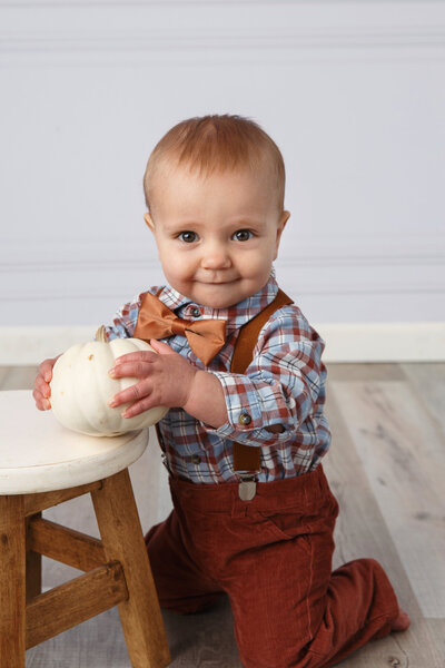 Portrait of a one year old boy kneeling next to a stool and holding a white pumpkin