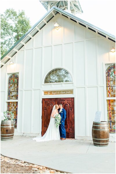 bride and groom kissing in front of white chapel with stained glass windows holding a white floral bouquet and greenery photographed by greenville wedding photographer