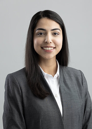 woman with straight dark hair and a gray suit smiling at the camera with a gray backdrop behind her.
