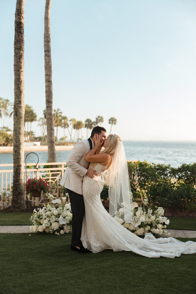 Bride and groom share a romantic kiss on the waterfront lawn at The Ritz-Carlton Sarasota, Florida—an intimate and elegant wedding moment at a luxury coastal venue, planned by Watters & Co. Events.