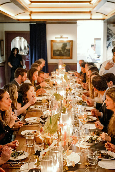 Wedding guests gathered around a long candlelit table, sharing a meal and toasting in a warmly lit dining space.