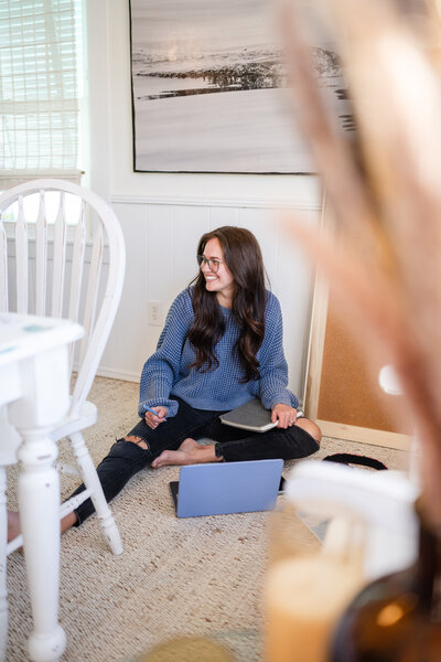 woman sits on the floor with pen and notebook in hands looking left while wearing a blue sweater, glasses, and a happy smile in wilmington, north carolina