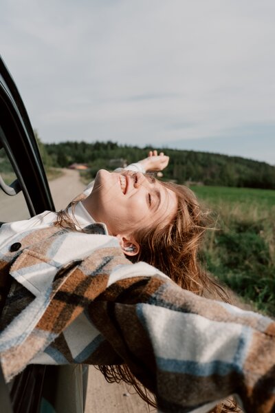Woman with red hair wearing flannel leans out of car window and spreads her arms while smiling with her eyes closed.