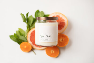 A white Citrus + Basil fragrance candle sitting on top of orange slices and basil leaves on a white backdrop
