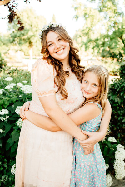 Colorful image of 2 girls embracing.  They are in a beautiful rose garden, wearing spring dresses. 
