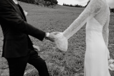 Two people in a black suit and white dress hold hands in front of wooded hill