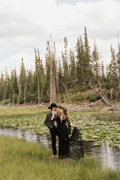 a bride and groom standing next to a lake while looking at each other with a smile