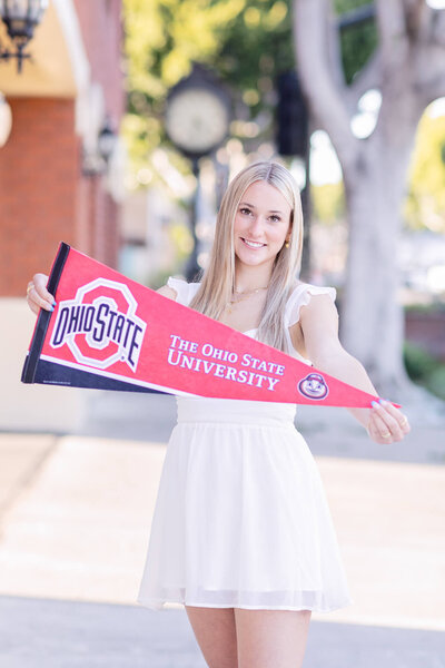 A blonde young woman wearing a white dress holding an Ohio State University banner smiling at the camera  in Old Town Tustin