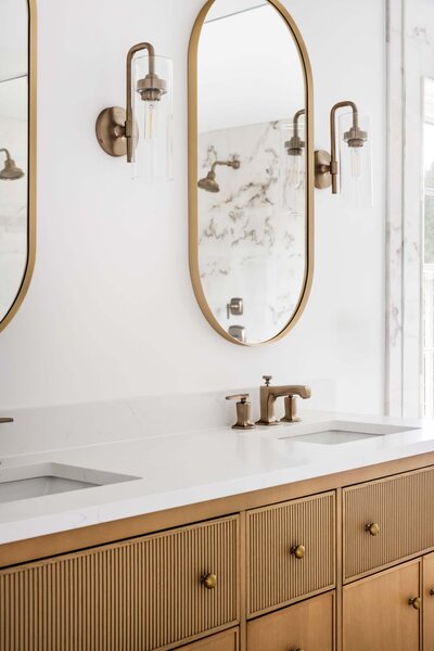 Modern bathroom vanity with ribbed wood cabinetry, white quartz countertop, brass fixtures, and oval brass-framed mirrors.