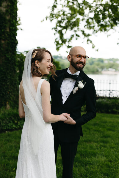 The bride and groom looking at their friends who are cheering for them during their couple photos. They're holding hands with the Susquehanna River behind them. 