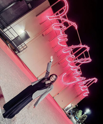 The Merry Hay in black jumpsuit and blazer in front of pink neon sign that reads "you belong here"