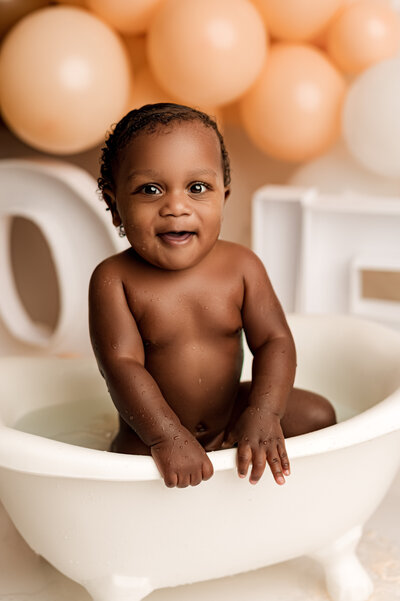 One year old baby girl playing in a mini bath slashing the water while looking into the camera during her cake smash session at NicSo Studio.