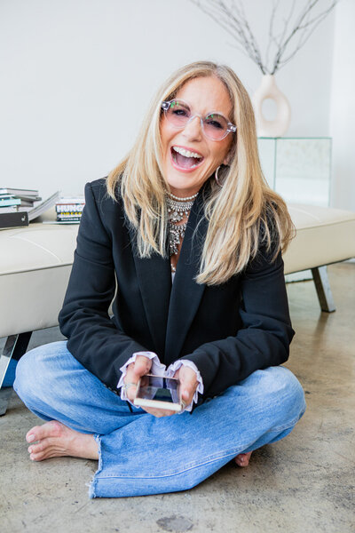 woman sitting cross-legged on the floor smiling and holding a phone while wearing a black blazer and jeans