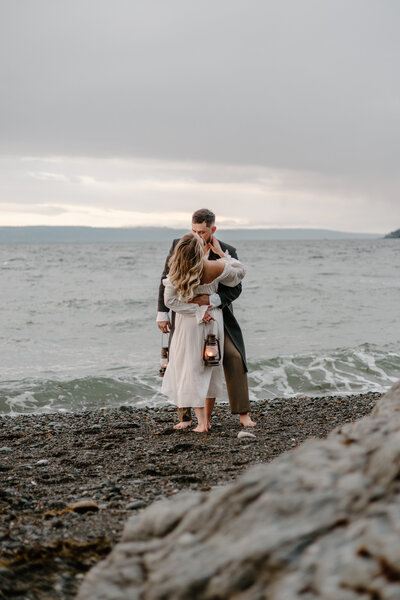Couple embraces on Seldovia beach with bride in white dress and lantern.